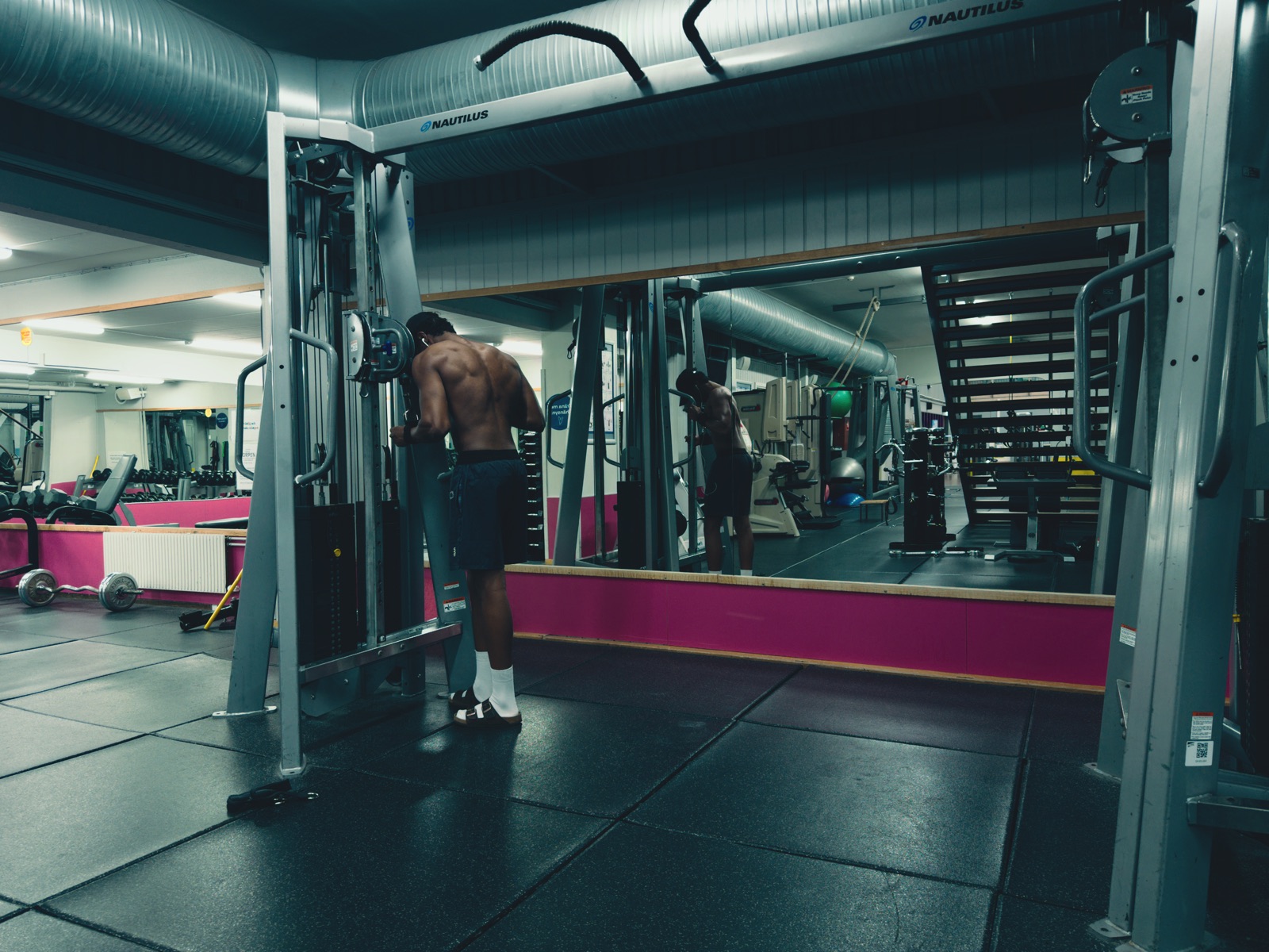 A man training on a cable machine in a gym with pink trim, his reflection in the mirror behind him