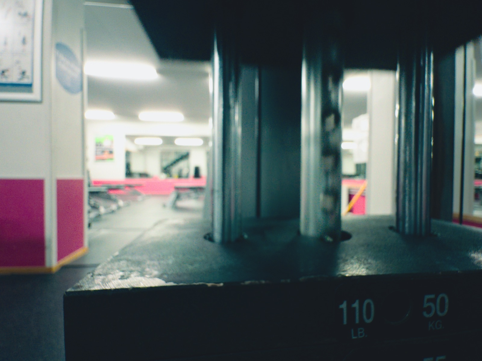 Close-up of a weight stack plate marked 110 lb and 50 kg, with a bright gym out of focus behind it